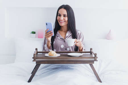 Photo of charming sweet woman nightwear sitting bed enjoying morning coffee reading modern gadget inside indoors home roomの写真素材