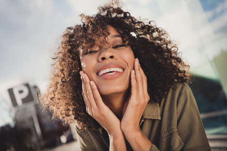 Portrait of adorable curly hairstyle dark skin lady hands on cheeks toothy beaming smile enjoy spending time outdoorsの写真素材