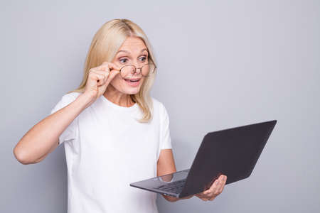 Photo portrait of amazed old woman taking off glasses holding laptop in one hand isolated on grey colored backgroundの写真素材