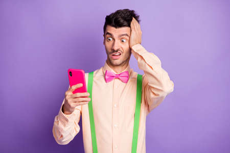 Photo of shocked astonished brunette man hold hand head look read fake news phone isolated on purple...