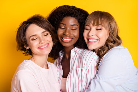 Portrait of three attractive dreamy affectionate cheerful girls bonding embracing isolated over bright yellow color backgroundの写真素材