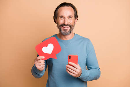Photo portrait of man holding phone red like heart card in hands isolated on pastel beige colored...