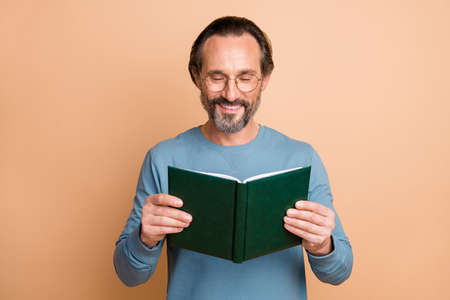 Photo of cheerful clever man hold textbook read wear eyeglasses blue pullover isolated beige color backgroundの写真素材