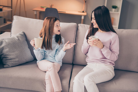Portrait of attractive cheerful girls daughter mommy sitting on cosy divan talking drinking cacao at house flat indoorの写真素材