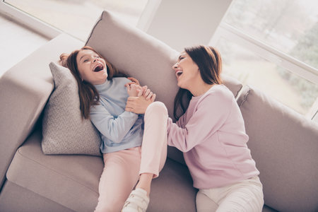 Portrait of attractive ecstatic cheerful girls daughter mommy lying on cozy divan tickling having fun at light house flat indoorの写真素材