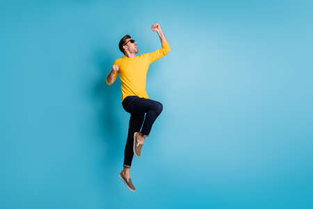 Full length profile portrait of delighted guy jump fists up open mouth shout yes isolated on blue color backgroundの写真素材