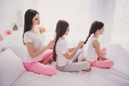 Photo of sweet attractive three siblings nightwear sitting bed braiding brushing hair indoors inside room homeの写真素材