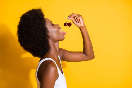 Close-up profile side view portrait of charming funky wavy-haired girl pulling cherry in mouth isolated on bright yellow color backgroundの写真素材