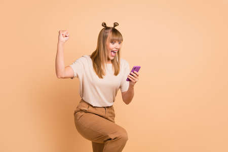 Photo of excited crazy girl hold smartphone raise fist scream open mouth wear t-shirt isolated beige color backgroundの写真素材