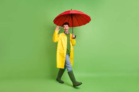 Full size photo of brunet impressed guy go wave hand with umbrella wear cloak boots isolated on green color backgroundの写真素材