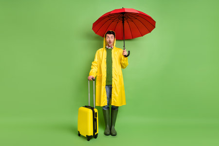 Full size photo of young man amazed hold umbrella wear raincoat baggage trip tourism flight isolated over green color backgroundの写真素材