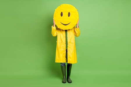 Full body photo of young man close cover face paper pinata emoji smile wear raincoat isolated over green color backgroundの写真素材