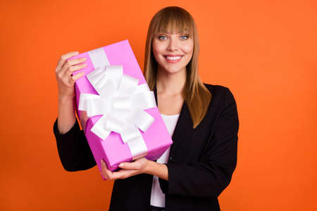 Portrait of charming cheerful girl holding in hands festal giftbox isolated over bright orange color backgroundの写真素材
