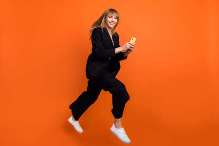 Full size photo of happy cheerful smiling good mood woman jumping using cellphone isolated on orange color backgroundの写真素材