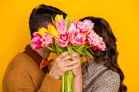 Photo of charming man and woman hold hands flowers cover close face kiss isolated on yellow color backgroundの写真素材