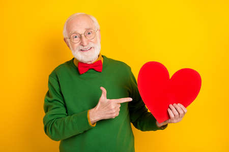 Photo of dreamy pretty mature man dressed pullover bow tie glasses pointing finger red heart isolated yellow color backgroundの写真素材