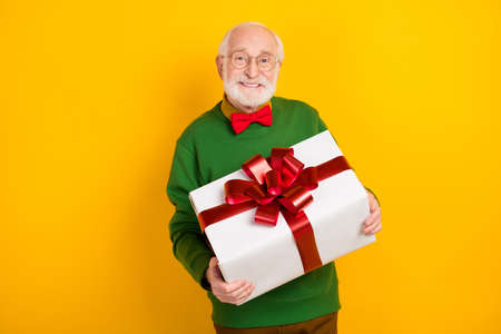 Photo of pretty positive mature man dressed pullover bow tie glasses holding big gift box isolated yellow color backgroundの写真素材