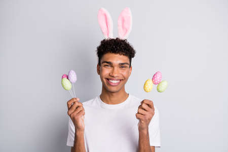 Photo of brunet optimistic curly guy hold eggs wear white t-shirt rabbit ears isolated on grey color...