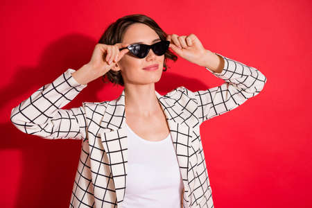 Photo of young attractive woman happy positive smile look empty space hands touch sunglasses isolated over red color backgroundの写真素材