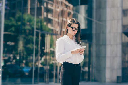 Photo of young attractive beautiful charming serious businesswoman in glasses typing in smartphone outside outdoorsの写真素材