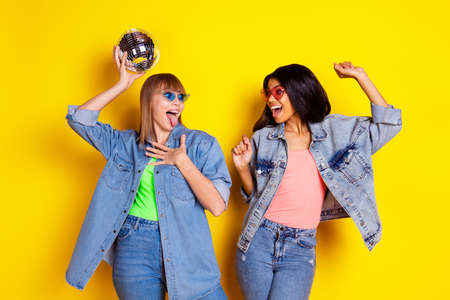 Portrait of two carefree girls arm hold disco ball open mouth tongue out have fun isolated on yellow color backgroundの写真素材