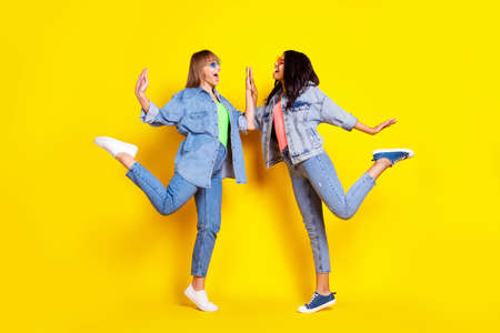 Full body profile photo of carefree excited pretty ladies look each other arms high five isolated on yellow color backgroundの写真素材
