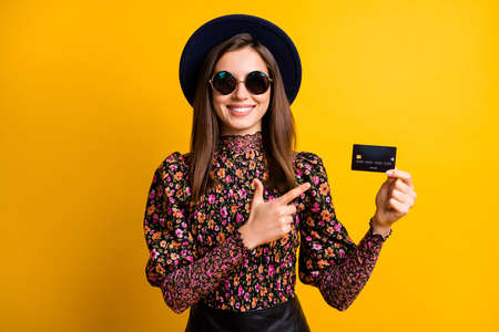 Photo portrait of stylish girl in headwear pointing finger credit card smiling in sunglass isolated on vivid yellow color backgroundの写真素材