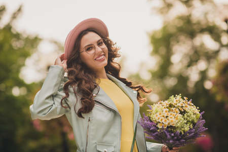 Portrait of pretty positive lady arm touch cap hold wild flowers look camera beaming smile weekend...