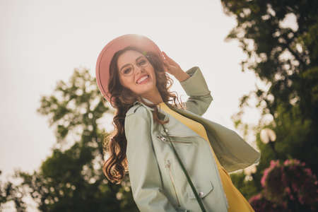 Photo of cute charming young lady wear turquoise jacket spectacles hand headwear smiling walking outside city parkの写真素材