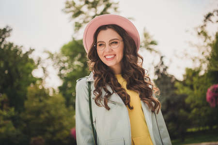 Photo of charming sweet young lady wear turquoise jacket headwear spectacles smiling walking outside city parkの写真素材