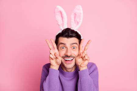 Photo of charming funky young man dressed purple pullover rabbit ears showing v-signs cheeks isolated pink color backgroundの写真素材