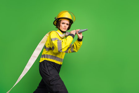 Photo of confident furious young fire lady workwear yellow helmet holding water hose isolated green color backgroundの写真素材