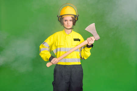 Photo of pretty serious young firewoman dressed yellow uniform helmet holding hatchet isolated green color backgroundの写真素材