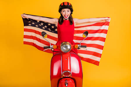 Photo of excited young lady wear red dress headwear sitting retro bike holding American flag isolated yellow color backgroundの写真素材