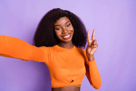 Photo of funny cute dark skin woman dressed orange shirt tacking selfie showing v-sign isolated purple color backgroundの写真素材