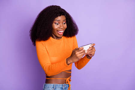 Photo of pretty excited dark skin lady wear orange shirt playing game modern device isolated violet...