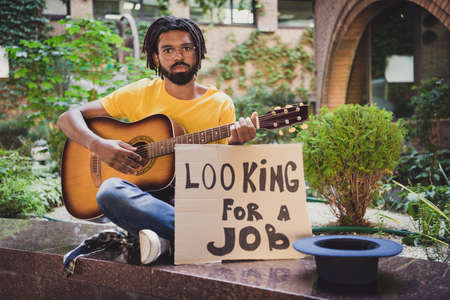 Photo portrait of guy playing guitar sitting sad on green street in summer looking for job need...