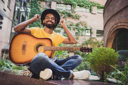 Photo portrait of man in black hat keeping guitar sitting on street in summer smiling