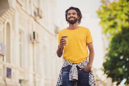 Photo portrait of young man wearing stylish outfit dreadlocks drinking coffee while walking on city streets with green treesの写真素材