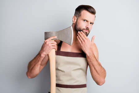 Portrait of attractive brutal guy wearing apron holding wooden ax touching beard isolated over light...