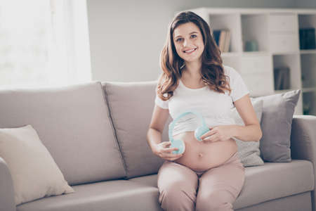 Photo of cheerful positive young woman sit couch living room hold headphone listen baby indoors inside houseの写真素材