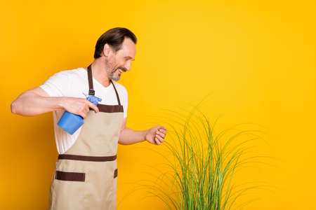 Profile photo of mature guy hold spray wash plant wear white t-shirt apron isolated yellow backgroundの写真素材
