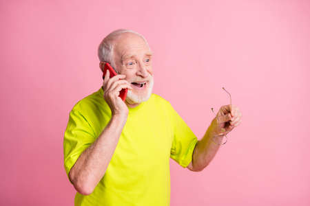 Photo of old cheerful positive man hold glasses wear lime t-shirt talk speak phone isolated on pink...
