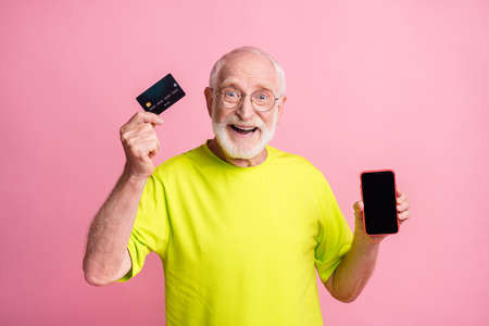 Photo of old excited cheerful smiling man wear lime t-shirt hold smartphone credit card isolated on...