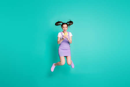 Full length body size photo of brunette girl with tails jumping high using smartphone social media isolated on vivid teal color backgroundの写真素材