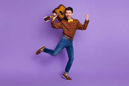Photo of funky crazy musician guy hold guitar dance wear bow tie plaid shirt jeans shoes isolated purple backgroundの写真素材