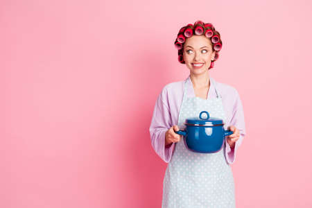 Photo of funny housewife dressed violet bathrobe red rollers holding saucepan looking empty space isolated pink color backgroundの写真素材