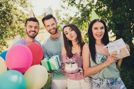 Photo of joyful brown haired happy friends at birthday party wear cones hold gift box cake balloons outside in picnic backyard outdoorsの写真素材