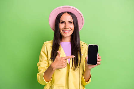 Photo of cute girl hold phone direct forefinger screen empty space wear yellow jacket pink cap isolated green color backgroundの写真素材