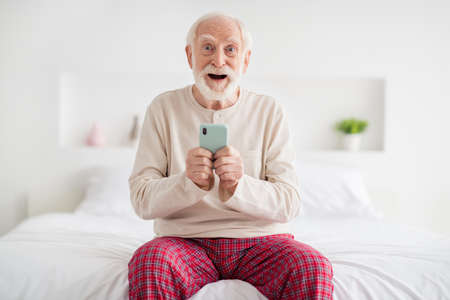 Photo of impressed funny mature man nightwear sitting bed typing modern device indoors inside room homeの写真素材